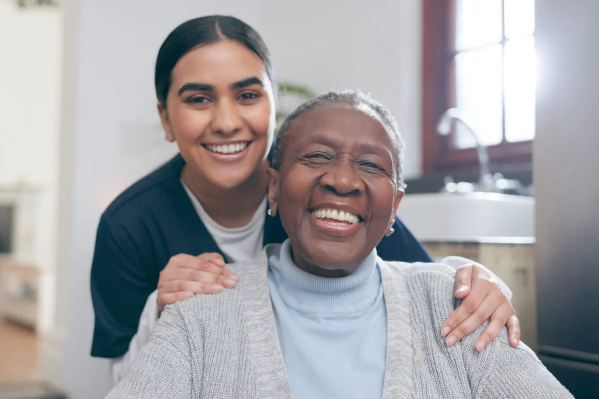 A smiling caregiver with her arm around an older woman at home