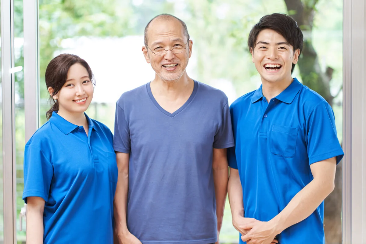 Two caregivers in blue uniforms standing with a smiling older man