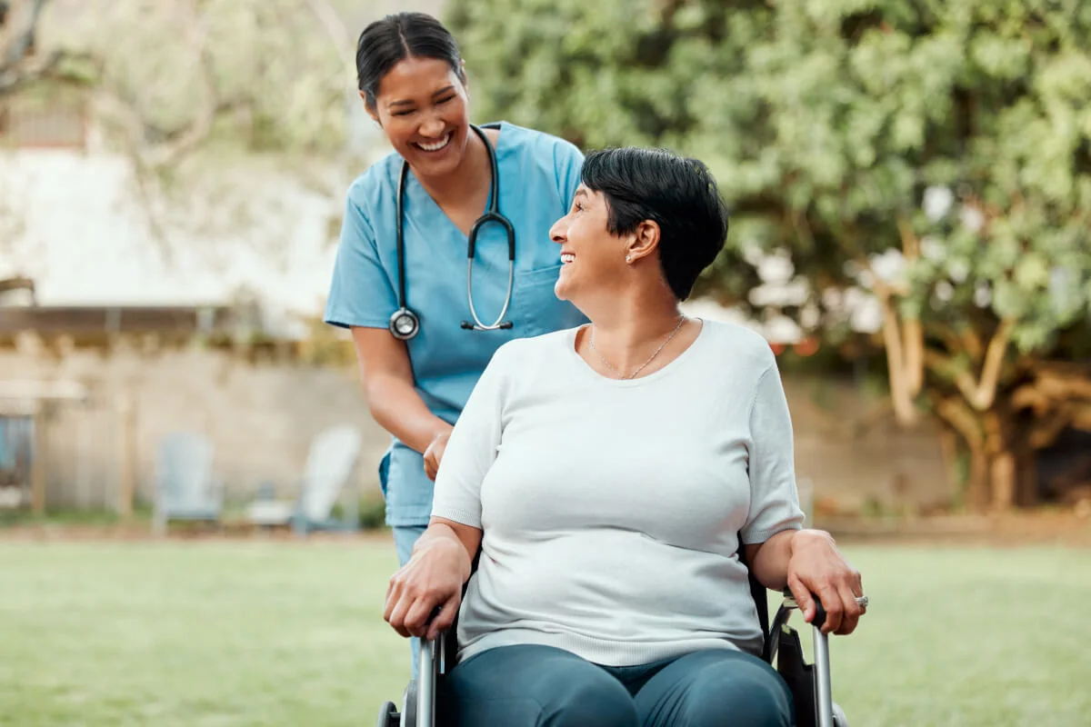 A nurse and a woman in a wheelchair laughing together outdoors